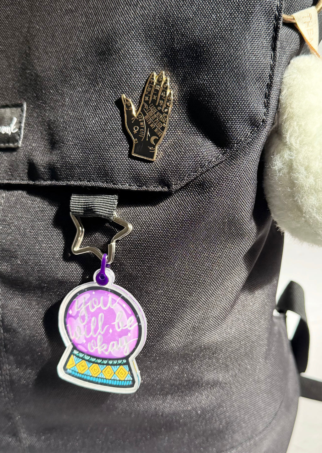 Backpack with a purple crystal ball charm and gold hand-shaped pin on a dark surface.