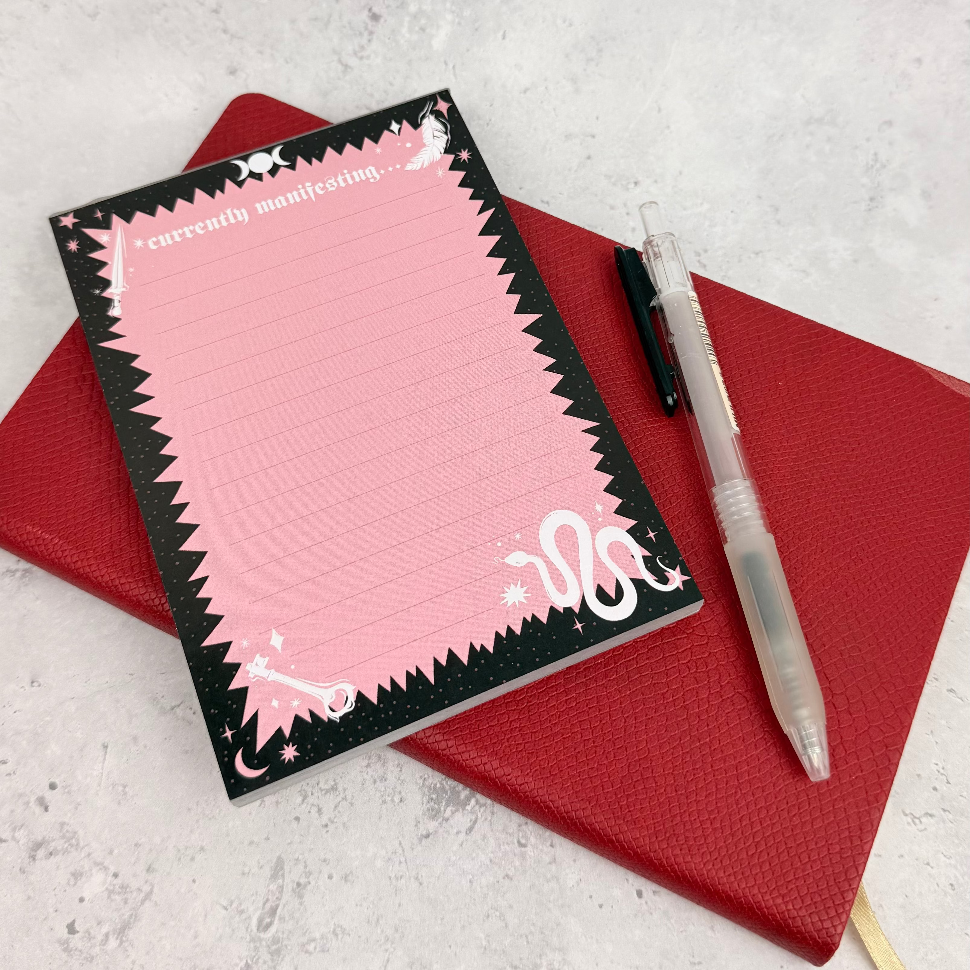 Pink notepad with black border on a red notebook with a pen on a light gray surface